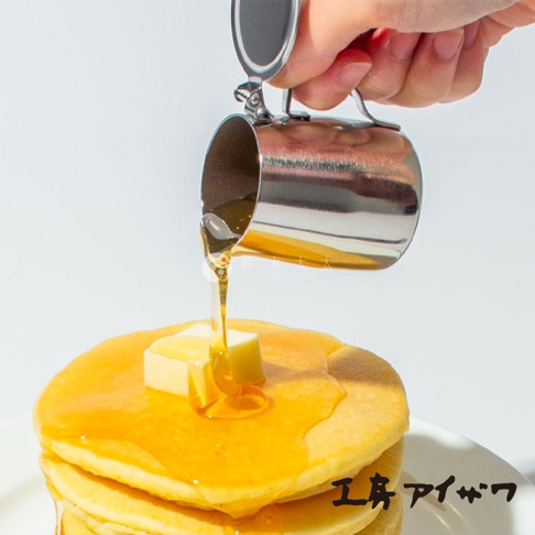 一個不鏽鋼附蓋蜂蜜盅，正將金黃色蜂蜜倒在一疊鬆餅和奶油上。