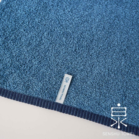 藍色長纖維毛巾，表面織有細緻的蓬鬆絨毛，邊緣飾有深藍色縫線。毛巾上印有「泉 SENSHU TOWEL」的品牌標誌。