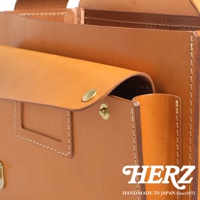 HERZ 手工皮革包，近拍展示其精緻縫線與金屬扣細節，品牌標誌與手工製造字樣清晰可見