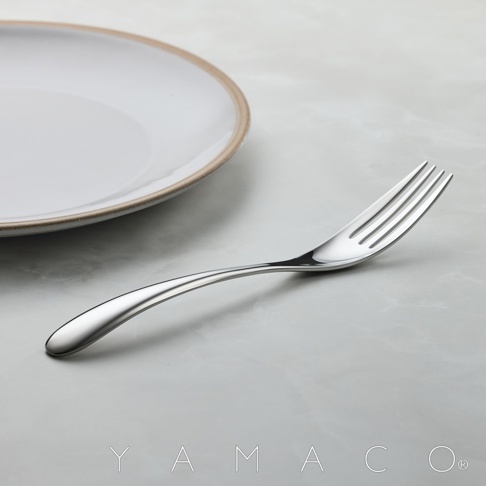 一個金屬叉子放在灰色大理石紋的桌面上，旁邊有一個白色的盤子，盤子的邊緣有淺棕色的裝飾。