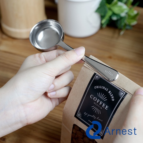 銀色不鏽鋼咖啡量匙夾，一端為圓形量匙，另一端為夾子，夾著印有「coffee」字樣的咖啡包。