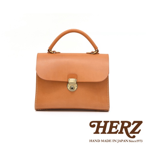 日本HERZ手工皮革包，焦糖色，翻蓋磁扣設計，頂部有提把，下方有HERZ品牌標誌與「HAND MADE IN JAPAN Since1973」字樣