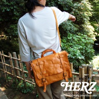 職人手作水洗加工皮革復古郵差包，以溫暖的橘棕色皮革製成，配有可調節的長肩帶、頂部提把，以及兩排帶有黃銅釦的皮帶裝飾，呈現經典郵差包造型。