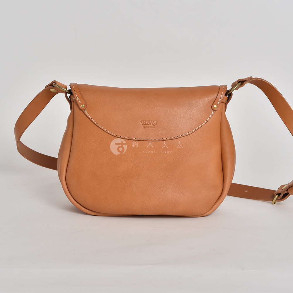 日本HERZ 職人手作皮革休閒旅行馬鞍包 CK-20，淺棕色皮革，以弧形縫線點綴，簡潔的品牌標誌與日文商家名稱置於包身中央，附有可調節長度的側背包帶。