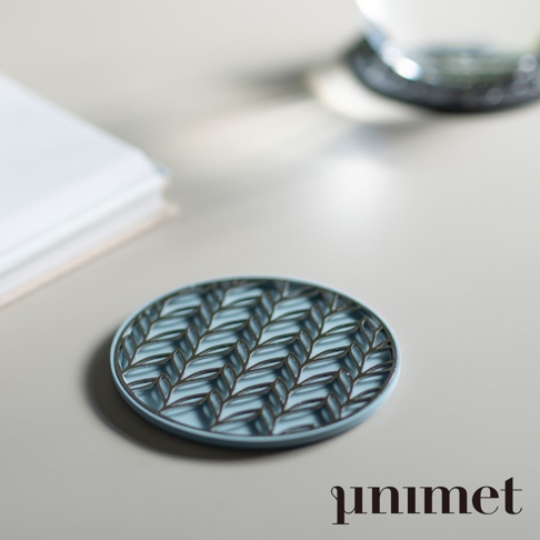 一張極具質感的 unimet 不鏽鋼光影藝術防滑杯墊特寫，圓形杯墊呈現柔和的淺藍色調，表面擁有精緻的鏤空葉脈紋理，在光線下投射出立體的交錯光影。杯墊擺放在簡約的桌面背景上，左側伴有一本筆記本，右上方則是一個模糊的玻璃杯底，整體呈現出靜謐且富有藝術氣息的居家氛圍。