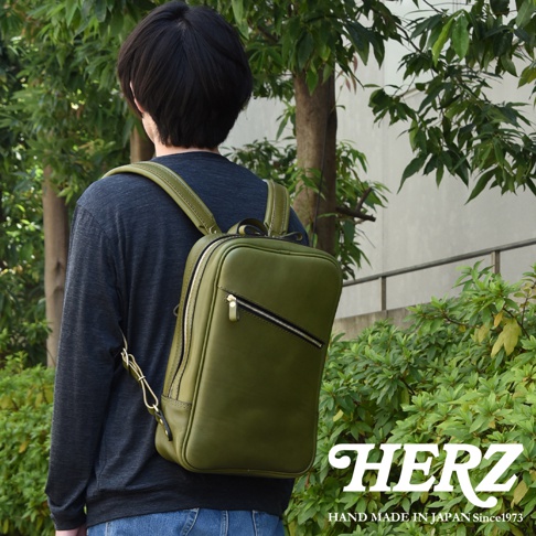 職人手作綠色皮革長方型多口袋後背包，展示於戶外。背包採用光滑皮革材質，設有拉鍊開合和外部拉鍊口袋。