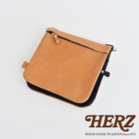 職人手作皮革翻轉雙色直式斜背包，以鞣製皮革製成，呈現自然的棕褐色調，搭配黑色包邊與內裡。包款設有拉鍊開口、皮質拉環，以及用於連接背帶的金屬環。