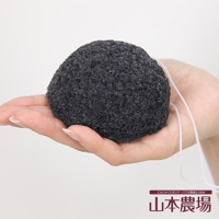 手持一顆深灰色、質地粗糙的蒟蒻洗臉海綿，海綿呈圓球狀，表面有許多細小孔洞，一條細白色掛繩從海綿後方延伸而出。