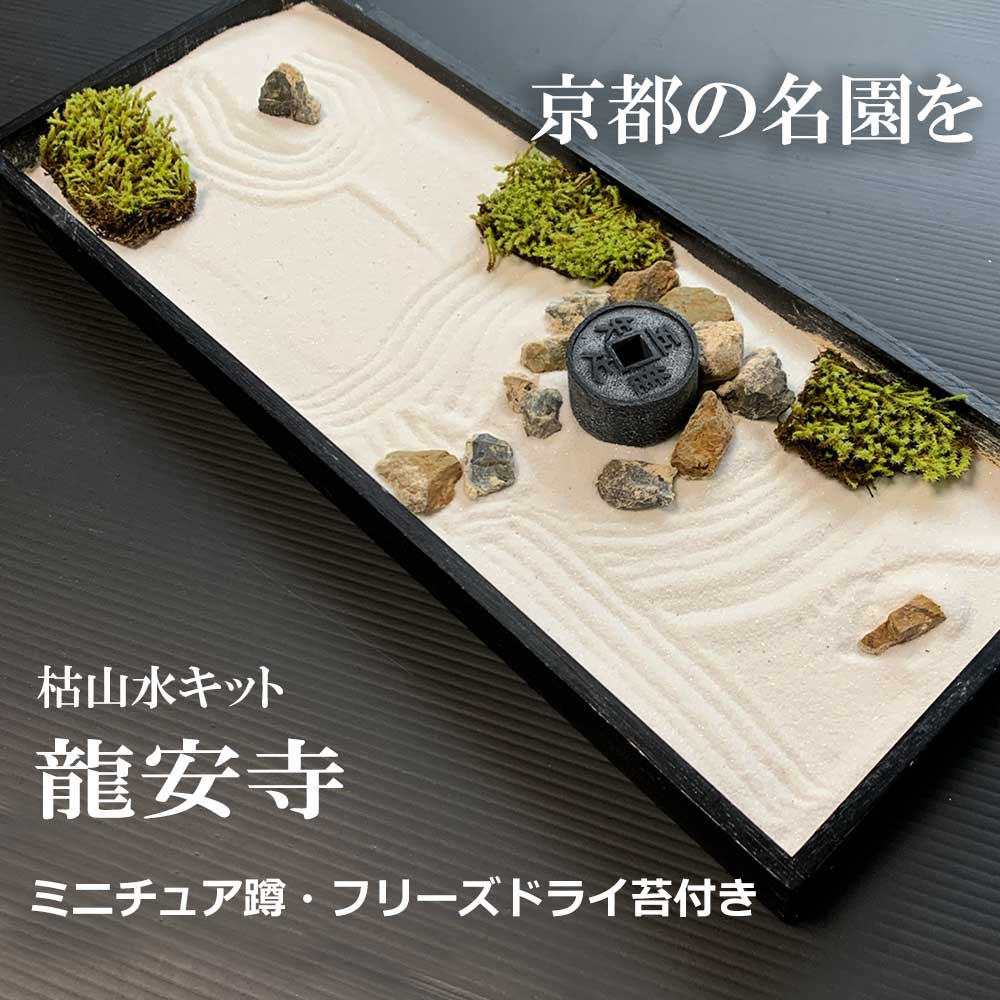 日本枯山水模型，以細膩的白色沙子鋪底，耙出波浪紋理。模型中點綴著幾塊灰褐色石塊，以及叢生的綠色苔蘚。中央有一個仿古錢幣造型的黑色裝飾物，整體呈現寧靜的日式庭園風格。