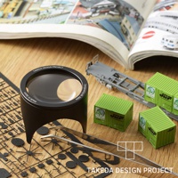 黑色鋁製閱讀放大鏡，鏡面印有「TAKEDA DESIGN PROJECT」及「Made in Japan」字樣，旁邊有綠色貨櫃模型、鐵路貨車模型及模型零件。