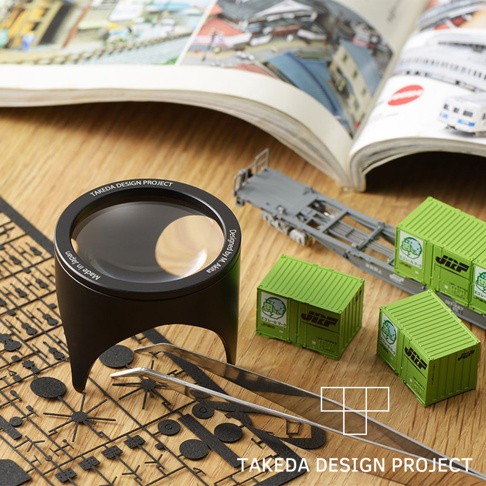 黑色鋁製閱讀放大鏡，鏡面印有「TAKEDA DESIGN PROJECT」及「Made in Japan」字樣，旁邊有綠色貨櫃模型、鐵路貨車模型及模型零件。