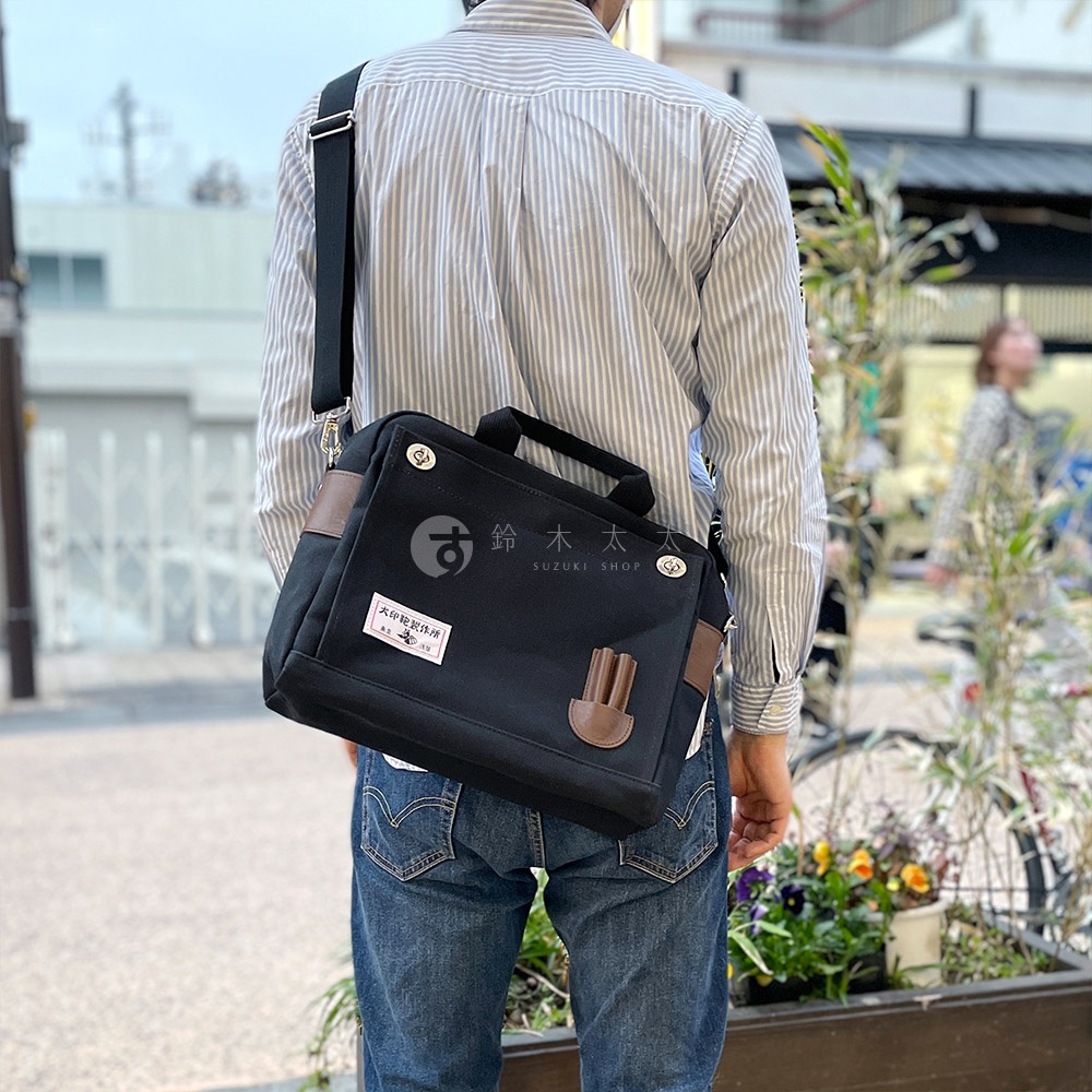 黑色純綿帆布製成的橫式側背包，配有咖啡色皮革裝飾，包包上有日文標籤和品牌標誌，可肩背或手提。