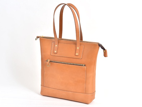 職人手作皮革威靈頓直式拉鍊托特包，天然皮革材質，淺棕色，配有頂部拉鍊開合、正面拉鍊口袋、兩個金屬鉚釘裝飾的皮片，以及一對長手提帶。
