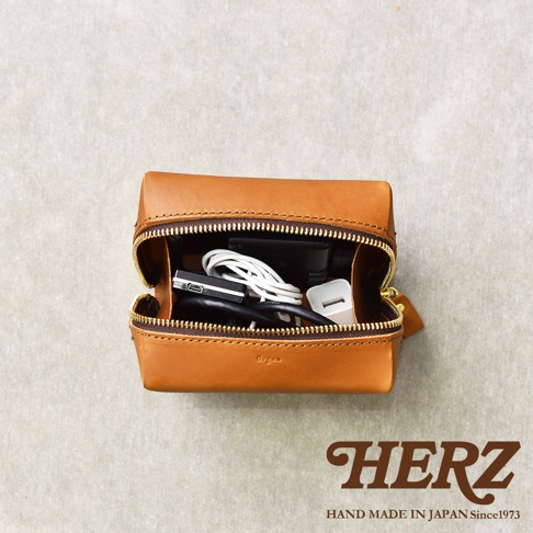 職人手作的棕色皮革Organ箱型收納包，拉鍊打開，內部裝有充電器、充電線等電子配件，上方印有HERZ品牌字樣。