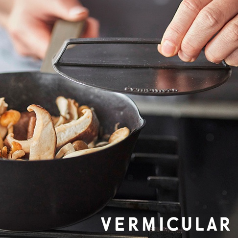 一隻手正拿著圓形的黑色 Vermicular 琺瑯鑄鐵落蓋，懸空置於正在烹調菇類的鑄鐵鍋上方，鍋中食材冒著熱氣，背景為深色的爐具。