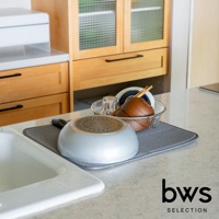 灰色可折疊收納瀝水墊上放置著一個銀色平底鍋，鍋底朝上。旁邊有一個金屬網籃，籃中裝有玻璃杯、木製碗和筷子。