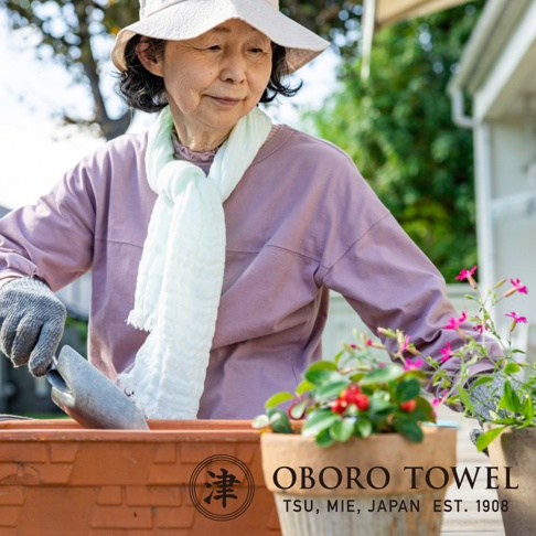 一位長者戴著帽子，身穿淡紫色長袖上衣，脖子上圍著一條淺綠色三層紗純棉掛脖毛巾，正在進行園藝活動。