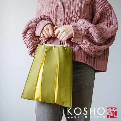一位女性穿著粉色針織衫搭配深色長褲，雙手提著一只由京都 KOSHO 設計的 ougi 帆布信玄袋，包款呈現獨特的橄欖綠色與黃色拼接百褶設計，搭配簡約的繩索手把，整體風格溫潤且具京都工藝質感。