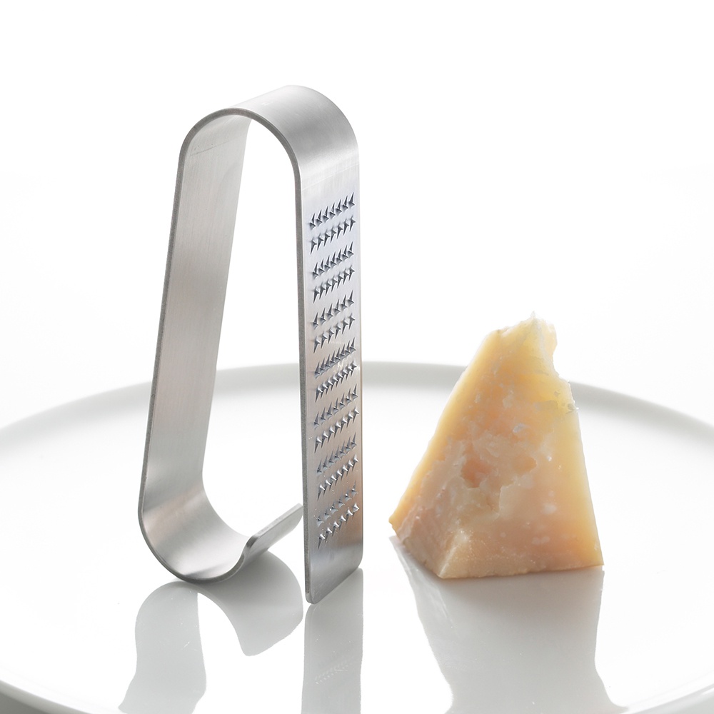 銀色不銹鋼立式磨泥器，帶有 U 型握柄和細密鋸齒狀研磨面，旁邊放著一塊淺黃色硬質起司。