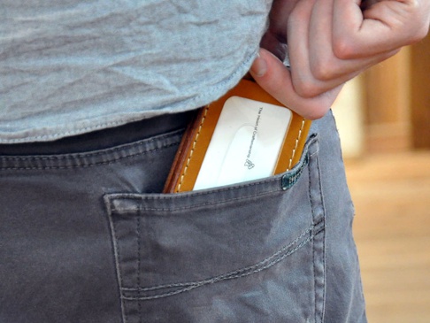 棕色皮革三面雙折票券收納夾，露出部分為其側面，內夾一張白色卡片，放入灰色褲子後口袋。