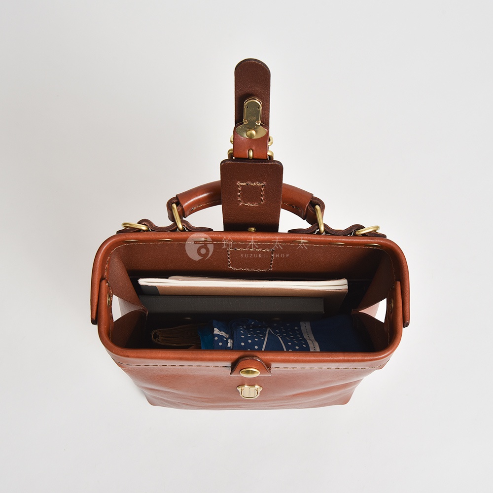職人手作皮革迷你軟皮杜勒斯包，棕色牛皮材質，開啟的包內裝有筆記本和摺疊傘，包身有金屬旋轉鎖扣和提把。
