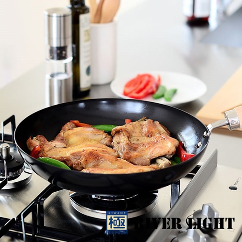 黑色不易生鏽平底鍋，鍋內有烹煮中的雞肉塊、青椒和紅椒，鍋柄為木質，置於瓦斯爐上。