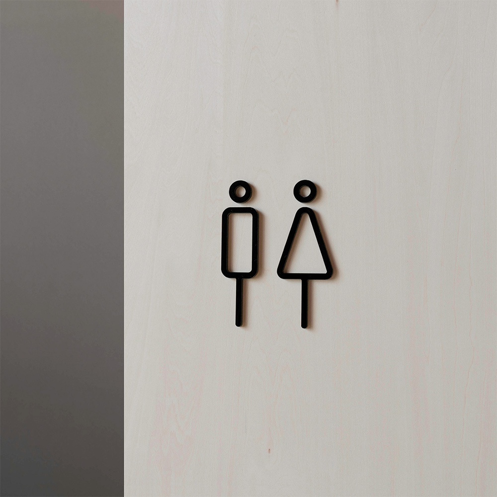 黑色線條勾勒的男女廁所標示牌，分別為方形身體的男性圖示和三角形身體的女性圖示，安裝於淺色木紋背景上。