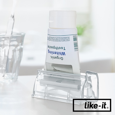 透明塑膠製的手動轉轉擠牙膏夾，夾著一支白色標有「Organic Whitening Toothpaste」字樣的牙膏，產品背景為淺色調，一旁有透明玻璃杯。