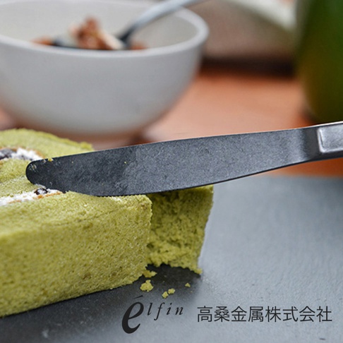 日製熔岩黑石餐刀，刀身呈現深邃的熔岩黑色，材質為金屬，表面帶有細緻的紋理。刀鋒正在切割綠色的蛋糕捲，後方為白色碗狀容器。