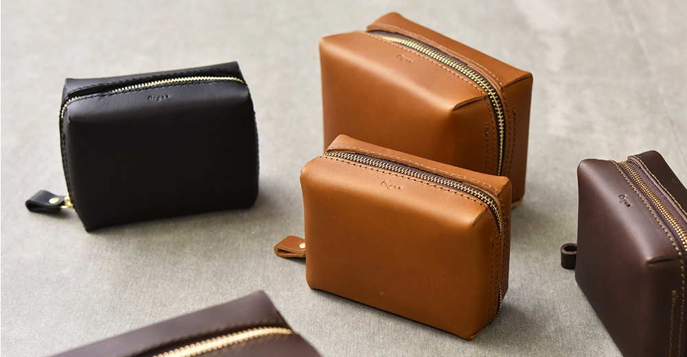 四個不同顏色（黑色、棕色、深棕色）的職人手作皮革Organ箱型收納包，以不同角度擺放在灰色背景上，展現其簡約的設計和細緻的皮革紋理。