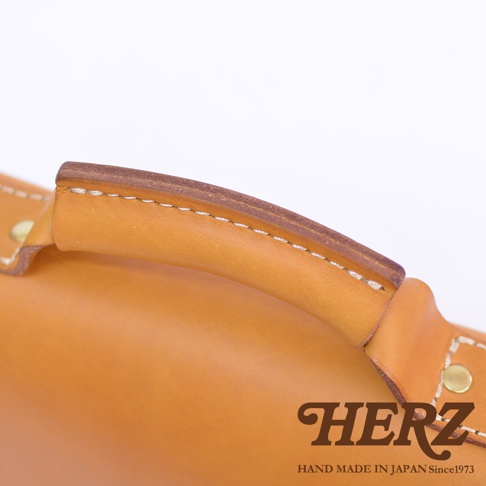 職人手作皮革經典兩用雙扣迷你單肩包，採用天然皮革製成，呈現溫潤的駝色調。包包的提把設計厚實，邊緣以白色縫線精緻縫合，展現細膩的工藝。金屬铆釘固定，增添復古質感。