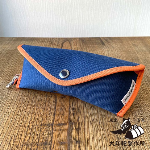 以犬印純綿帆布製作的藍色眼鏡收納盒，搭配橘色滾邊，正面有一個金屬扣，上面印有日文字樣。收納盒旁可見犬印鞄製作所的品牌標誌，包含黑色的狗剪影和一個行李箱，以及日文公司名稱。