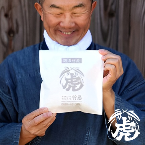 一位身穿藍色日式服飾的亞洲男性，雙手拿著一個白色包裝袋，上面印有銀色的「虎」字圖案和日文標示。