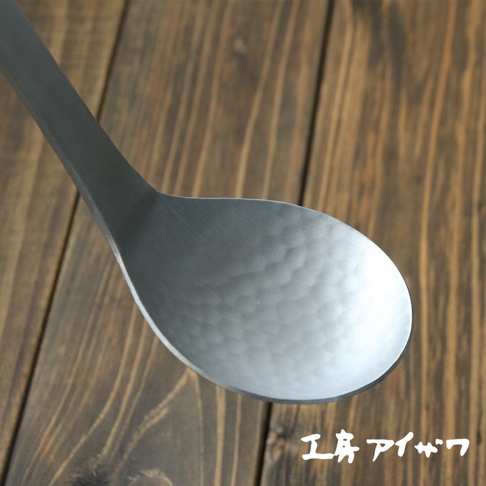 特寫鋁製湯勺，勺面有錘目紋，呈現銀灰色，勺柄連接勺面，背景為木紋桌面。