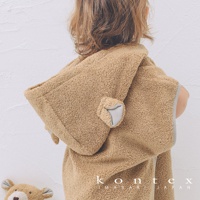 孩童穿著淺棕色毛絨材質的斗篷式連帽浴巾，浴巾上有可愛的熊耳朵設計，帽子內襯為淺灰色。