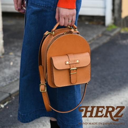 職人手作皮革迷你後背包，採用原色皮革，圓弧形設計，正面有一個帶有皮帶釦的翻蓋小口袋，頂部有提把，可肩背或手提。
