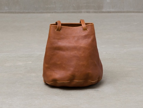 一個棕色皮革抽繩包，有兩個短提把，底部有波浪邊設計，放置在灰色背景上。