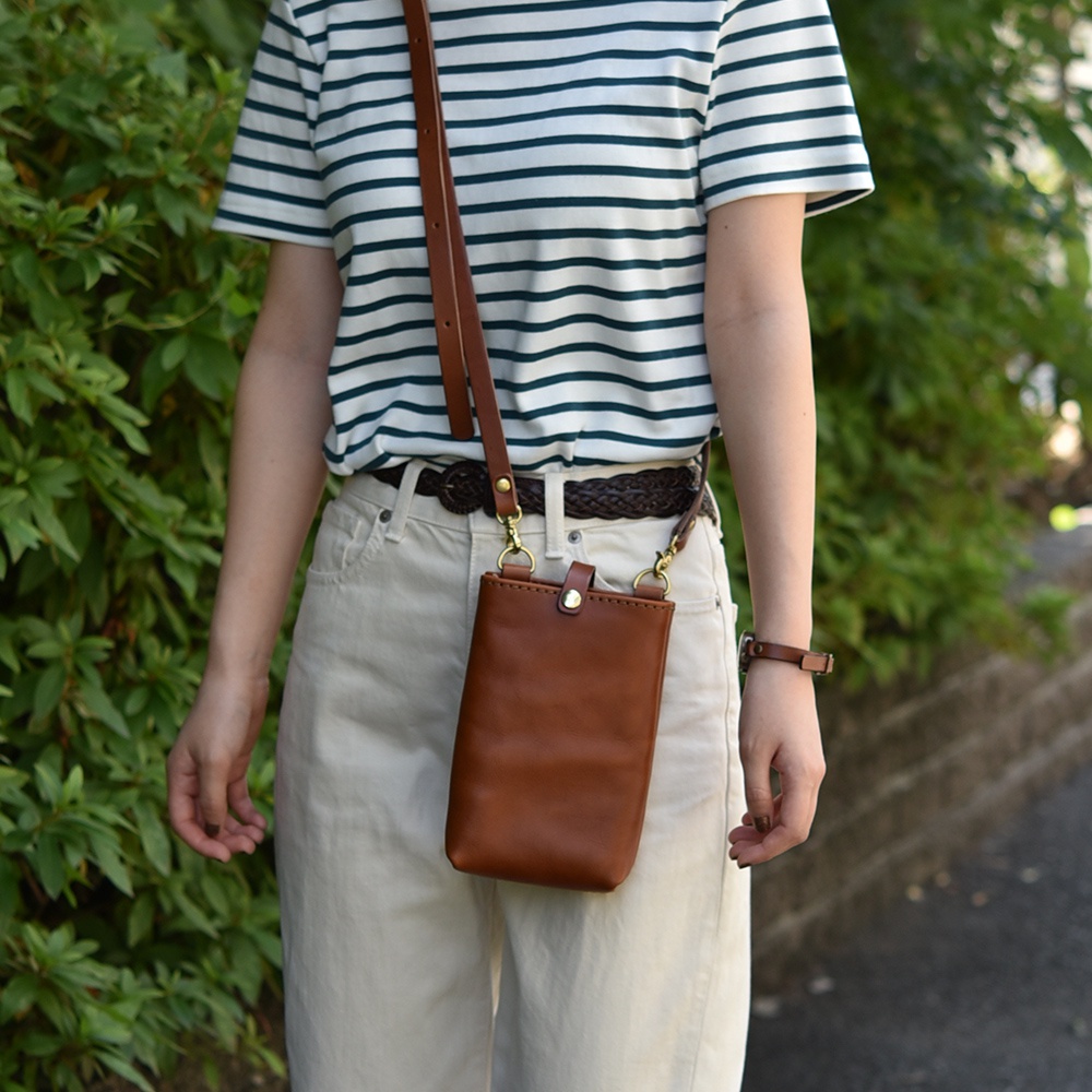 一位女性穿著條紋上衣和米色褲子，斜挎著一個棕褐色皮革手機包。包包上有金屬扣環和可調節的長肩帶。