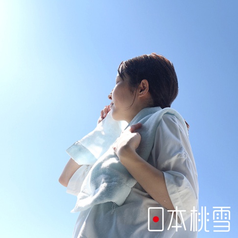 一位女性用淺藍色毛巾擦拭臉頰，毛巾掛在頸部，背景為晴朗的藍天。