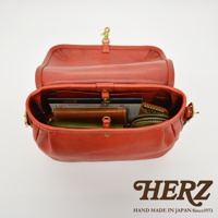 打開的橘紅色皮革復古翻蓋郵差包，內有筆記本、錢包、零錢包等多樣物品，包身線條圓潤，採用金屬扣環和縫線工藝。