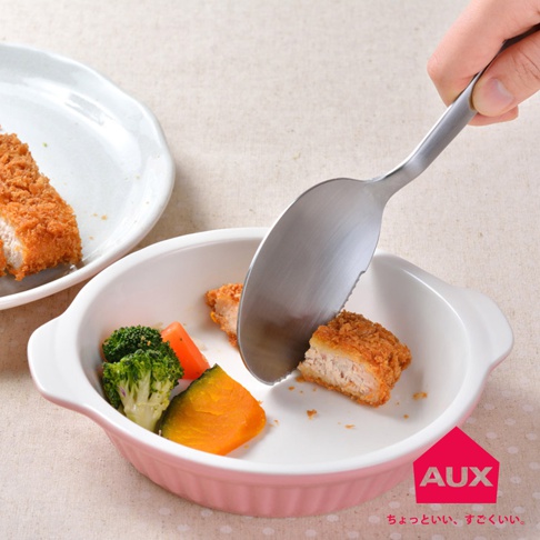 不鏽鋼切切分食匙，匙緣有鋸齒，正用來分切炸豬排，旁有花椰菜、紅蘿蔔塊與南瓜塊，盛裝於粉色陶瓷烤皿中。