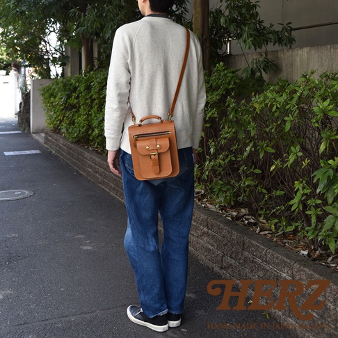 一名男子斜挎著一個棕色皮革兩用單肩包，包上有一個立體前袋，配有翻蓋和金屬釦。