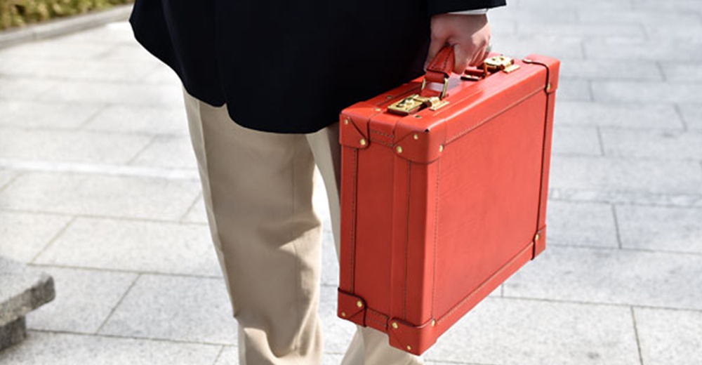 紅色皮革英倫風手提行李箱，設有金色扣環和金屬邊角裝飾，由人手提起。