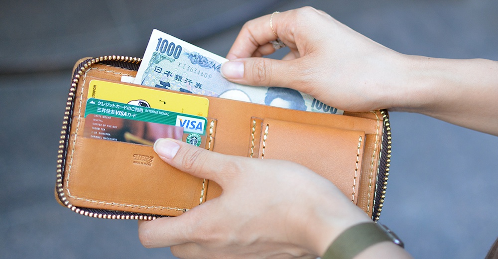 一位女性手持日本HERZ職人手作皮革拉鍊短夾，皮夾呈現溫潤的淺棕色皮革質感，內部夾層展示了信用卡及日圓紙鈔，畫面聚焦於手部與皮夾的細節互動。
