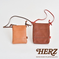 兩款職人手作皮革直式隨身單肩包，一款為淺棕色，另一款為深棕色，均採用光滑皮革材質，配有同色系或對比色的細長肩帶，包身正面有方形貼袋設計，並以白色車線縫合。