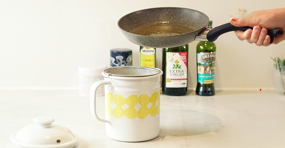 白色琺瑯濾油壺，壺身有黃色網格圖案，壺口上方放有金屬濾網。一隻手正從灰色平底鍋中傾倒液體。背景有數瓶橄欖油。