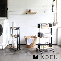 黑色金屬三層置物夾縫式收納推車，配有木質層板，可移動，有兩個置物架，其中一個架子上有白色瓶狀物，另一個架子上有疊好的白色毛巾。