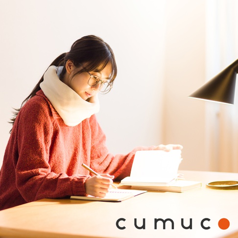 一位女性戴著保暖頸套，身穿紅色毛衣，戴著眼鏡，正在低頭在筆記本上寫字，桌上還有打開的書和檯燈，底部有CUMUCO的品牌標誌
