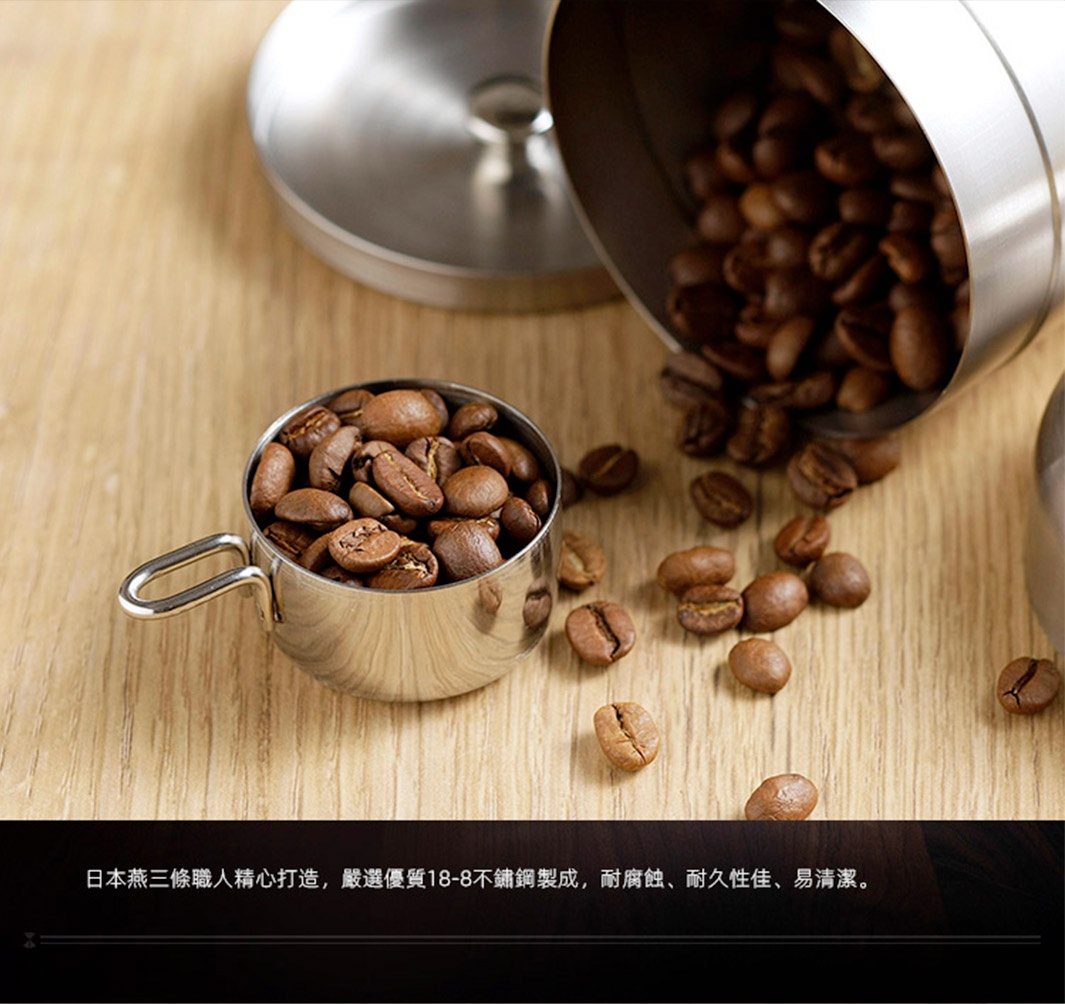 日製18-8不鏽鋼儲豆罐，附有銀色量匙，罐內及周圍散落著烘焙過的咖啡豆。