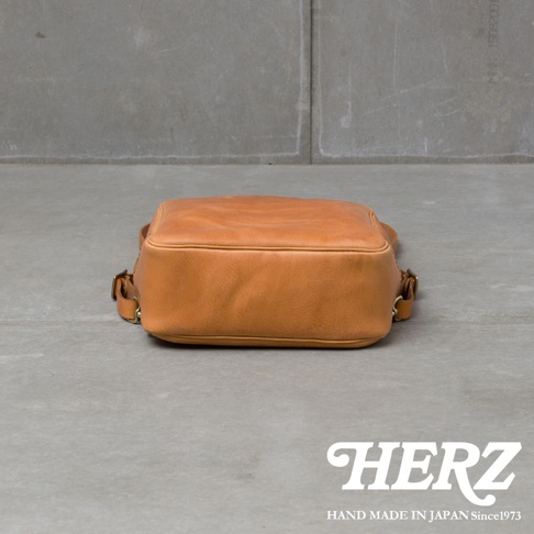 一個棕褐色皮革後背包，底部朝上，置於灰色水泥地面上，背景為灰色水泥牆。包包上有金屬扣環，底部印有HERZ字樣及HAND MADE IN JAPAN Since1973。